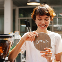 Young woman in coffee shop holding a wooden "open" sign in shape of speech bubble.